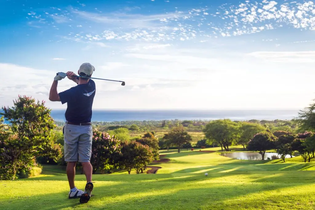 A golfer follows through on a drive above a green course with the ocean beyond